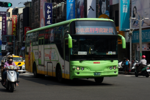 Ein grüner Bus fährt eine von hohen Gebäuden gesäumte Straße entlang, mit Menschen auf Motorrädern und anderen Fahrzeugen, einem Verkehrszeichen auf der linken Seite und Werbetafeln im Hintergrund unter einem klaren blauen Himmel.