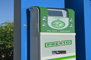 Ein grün-weißer Parkscheinautomat mit Knöpfen und Text auf einer blauen Wand, mit Bäumen und einem klaren blauen Himmel im Hintergrund.