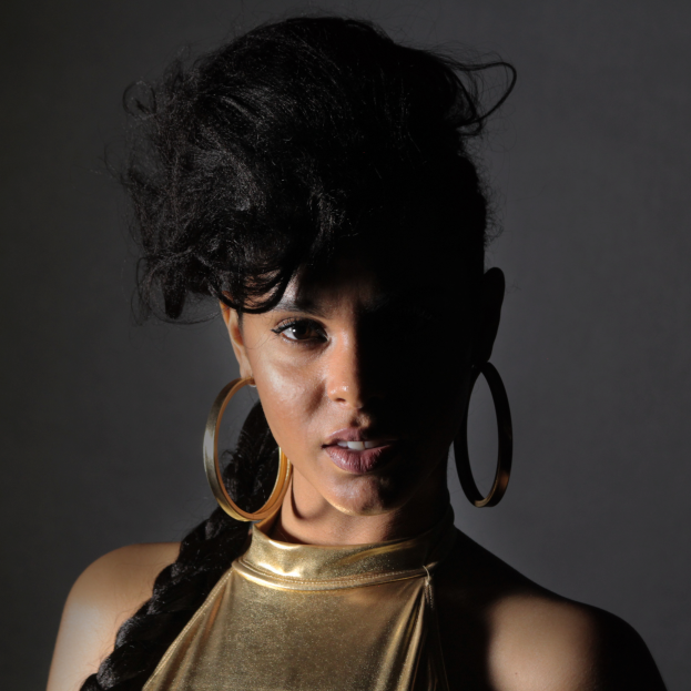 Eine Frau in einem goldenen Kleid und großen Ohrringen steht selbstbewusst vor einer Wand, ihre Haare sind glatt geschnitten und sie trägt ein dezentes, natürliches Make-up mit pinkem Rouge und glänzenden Lippen, blickt direkt in die Kamera mit einem entschlossenen Gesichtsausdruck.