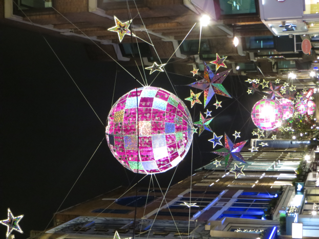 Eine Stadtstraße bei Nacht mit zahlreichen Lichtern und Dekorationen, darunter eine große Diskokugel, die von einem Gebäude hängt und eine festliche Atmosphäre schafft.