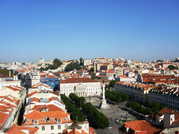 Blick auf Lissabon von einem Hügel aus, der Gebäude mit Fenstern, Bäume, eine Statue auf einem Sockel und einige Menschen auf der Straße zeigt, mit dem Himmel im Hintergrund.
