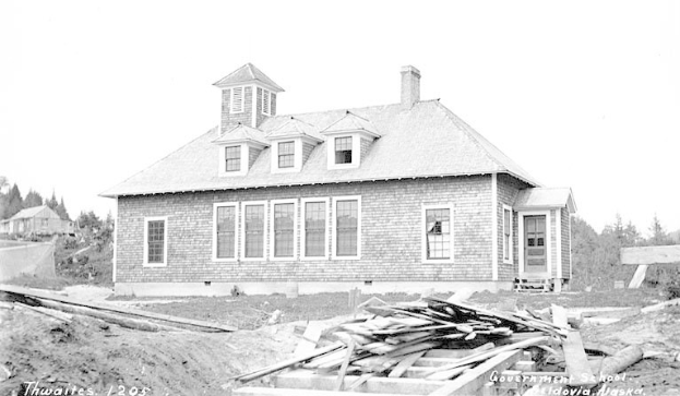 Schwarzes und weißes Foto eines zweistöckigen Gebäudes mit mehreren Fenstern und Türen, umgeben von Bäumen unter einem klaren Himmel, mit hölzernen Brettern am Boden, die "Regierungs-Schule, Thwaites, 1905" lesen.