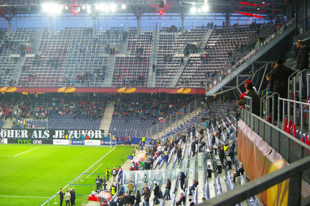 Großes Stadion mit Zuschauern bei einem Fußballspiel, einige stehen und sitzen auf Stühlen, eine Reling und ein Banner auf der rechten Seite und ein leerer Abschnitt mit Deckenleuchten im Hintergrund.