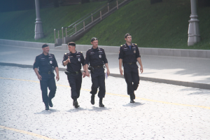 Drei Polizeibeamte in Uniform und Mützen, die eine Straße mit Pfählen, Geländern, Treppen und Gras im Hintergrund entlanggehen.