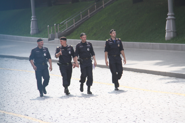 Drei Polizeibeamte in Uniform und Mützen, die eine Straße mit Pfählen, Geländern, Treppen und Gras im Hintergrund entlanggehen.
