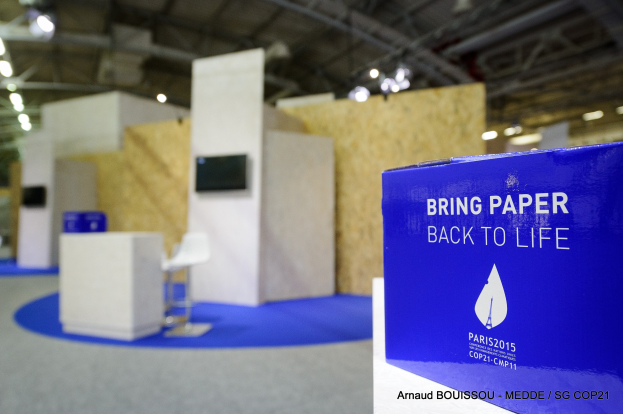 Messestand mit einem blauen und weißen "Bring Paper Back to Life"-Schild auf der rechten Seite, mit unscharfen Stühlen, Tischen, einem wandmontierten Fernseher und Deckenleuchten im Hintergrund.