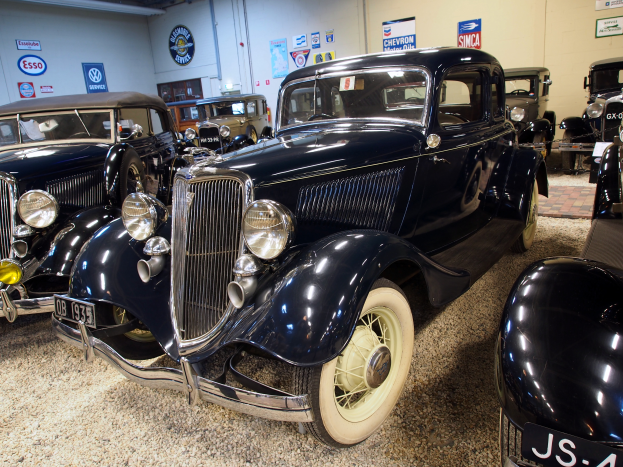 Eine Gruppe von Oldtimern in einer Garage geparkt, mit einer Wand im Hintergrund, die mit Brettern und Lampen an der Oberseite verziert ist, die den Eindruck eines klassischen Oldtimertreffens vermittelt.