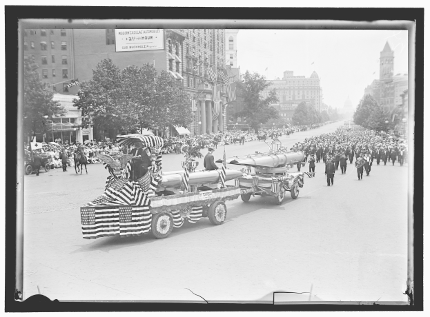 Schwarzes und weißes Foto eines Umzugs mit einem Feuerwehrauto in der Mitte, umgeben von Menschen, einige halten Fahnen, mit Bäumen und Gebäuden im Hintergrund und einer Tafel mit Text.