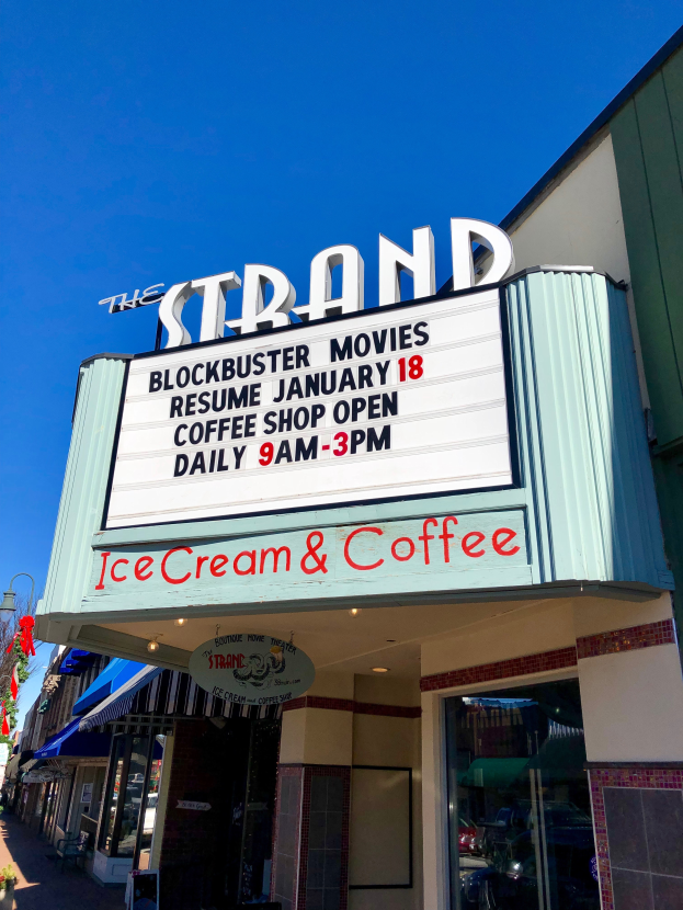 The Strand Blockbuster Movies Resume January 18 Coffee Shop building with a name board, a street pole, a group of trees, potted plants, and a cloudy sky.