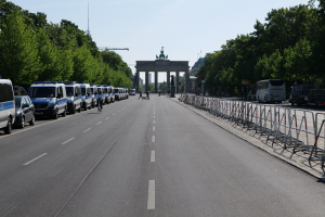Lange Reihe von Polizeiwagen auf der Seite einer Straße vor dem Brandenburger Tor in Berlin, Deutschland, mit Menschen auf Fahrrädern und in der Nähe, Barrieren, Bäumen, einem Bogen mit Statuen im Hintergrund und sichtbarem Himmel.