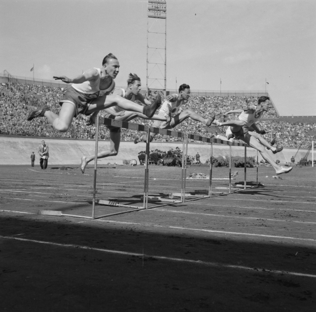 Eine Gruppe von Männern in Schwarz und Weiß springt über Hürden auf einer Laufbahn, im Hintergrund ist ein Stadion mit Zuschauern und in der Ferne ein Turm zu sehen.