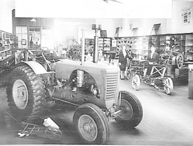 Ein Schwarz-Weiß-Foto eines Traktors in einer Fabrik mit mehreren Personen, anderen Fahrzeugen, einem Müllcontainer auf der rechten Seite und Regalen mit verschiedenen Gegenständen im Hintergrund.