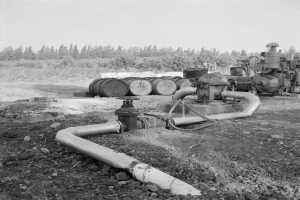 Ein Schwarz-Weiß-Foto einer Gasleitung in einem Feld, umgeben von Maschinen, Rohren und Tonnen, mit Bäumen und einem klaren Himmel im Hintergrund.