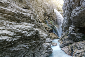 Kleiner Wasserfall, der über zerklüftete Felsen in einem steinigen Tal hinabstürzt, umgeben von saftig grünen Hügeln unter strahlendem Sonnenschein.