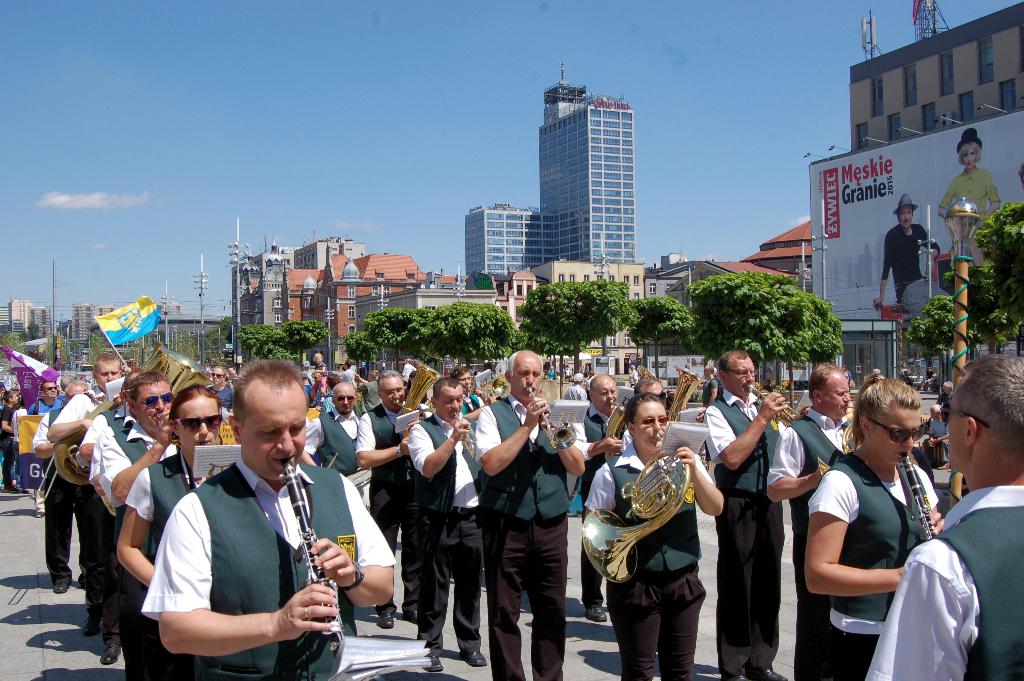Eine Gruppe von Menschen in einheitlichen Kostümen, die verschiedene Musikinstrumente spielen, draußen mit Bäumen, Pfählen, Fahnen und einem Banner mit Text und Bildern im Hintergrund.