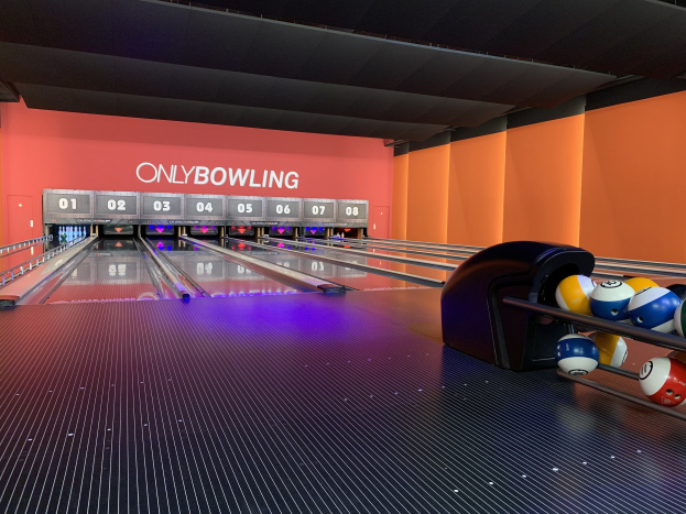 Eine Bowlingbahn mit Bowlingkugeln auf der rechten Seite, Bowlingpins in der Mitte und einer textbedeckten Wand im Hintergrund, mit der Decke oben.