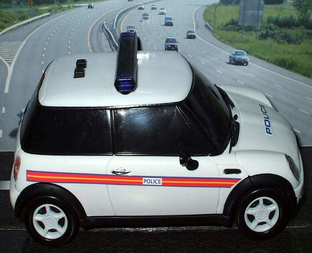 Weißer Polizeiwagen mit 'Polizei'-Aufschrift auf einer Straße außerhalb der Stadt, umgeben von Verkehr, Gras und Bäumen.