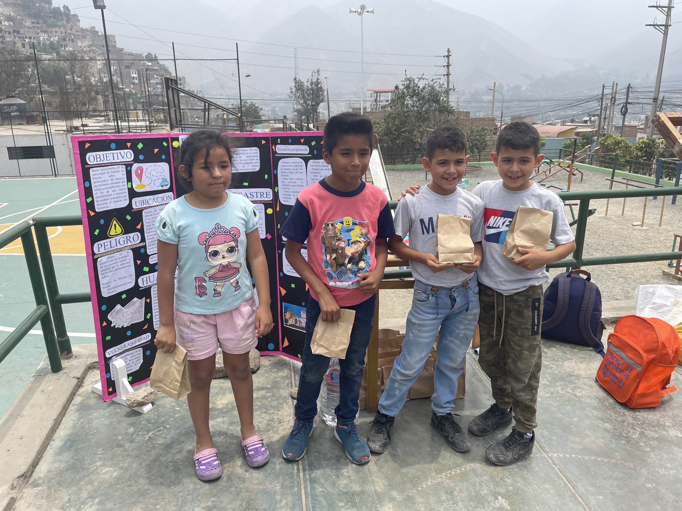 Gruppe von Kindern auf einem Basketballfeld mit Papiert├Ąten in den H├Ąnden, mit T├Ąten und Gegenst├Ąnden auf der rechten Seite und einer Tafel mit Text auf der linken Seite, vor einem Hintergrund aus Z├Ąunen, Masten, Stra├čenlaternen, B├Ąumen, Geb├Ąuden, H├Ąngen und Himmel.