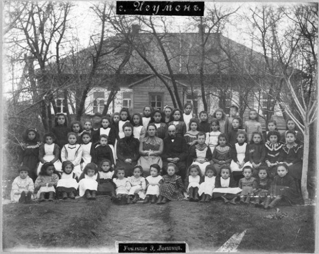 Schwarzes und weißes Foto einer Gruppe von Kindern, die vor einem Gebäude mit Fenstern posieren, umgeben von Bäumen und einem klaren Himmel, mit Text unten: "Jugoslawien, die erste Schule der Welt".