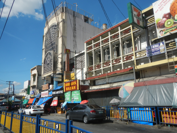 Eine belebte Stadtstraße mit Autos, hohen Gebäuden, Schildern, Regenschirmen, einem Metallzaun und Strommasten mit Drähten.