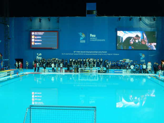 Gruppe von Menschen, die um ein Hallenbad mit Netz am Boden stehen, mit einer Wand im Hintergrund, die den Text und Logos 'FINA World Championships 2015 Kazan' anzeigt, sowie Gerüste und Lampen.