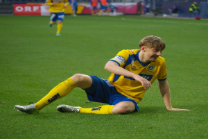 Ein Fußballspieler sitzt auf dem Boden und hat seinen Kopf in den Händen, umgeben von einer Gruppe von Menschen in Sportbekleidung, mit unscharfen Brettern, Geländern und Pfählen im Hintergrund.