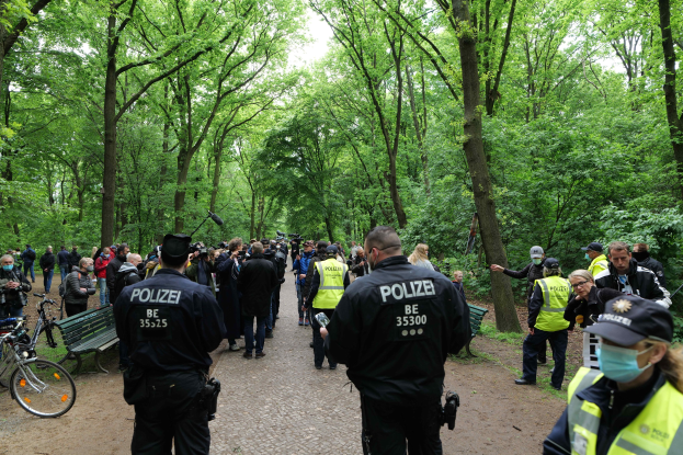 Eine Gruppe von Polizisten steht vor einer Menschenmenge, einige tragen Mützen und Masken, mit Fahrrädern und einer Bank im Vordergrund und Bäumen und Himmel im Hintergrund.