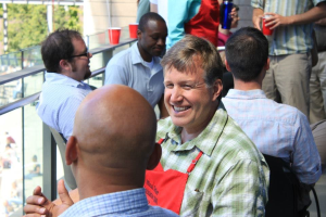 Chefkoch der Universität Texas in Austin in einer Schürze, der mit einem Mann auf einem Balkon spricht, umgeben von sitzenden und stehenden Zuhörern, Geländer auf der linken Seite, Bäume und ein Gebäude im Hintergrund.