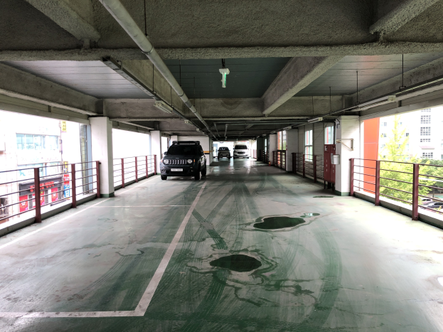 Ein mehrgeschossiges Parkhaus mit geparkten Autos, Geländern, Säulen, Deckenleuchten, Rohren und umgebenden Bäumen, vor einem Hintergrund von Gebäuden mit Fenstern in einer Stadtlandschaft.