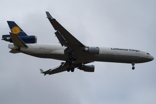Ein Lufthansa Cargo Boeing 777-300ER im Flug, mit ausgefahrenen Flügeln, aktiven Triebwerken und sichtbarem Text auf dem Rumpf gegen einen bewölkten Himmel.
