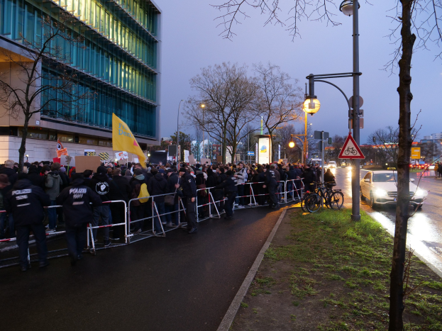 Eine große Gruppe von Menschen protestiert vor einem Gebäude in Berlin, hält Schilder, mit Barrikaden, Fahrrädern, Laternen, Schildern, Bäumen und Gras im Hintergrund.