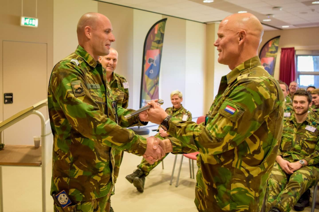 Zwei Männer in Militäruniformen geben sich die Hand vor einer sitzenden Gruppe, mit einem Tisch, Geländer, Fahnen, einem Schild, einem Fenster mit Vorhang und Deckenleuchten im Hintergrund, was auf eine deutsche Armee-Versammlung hinweist.