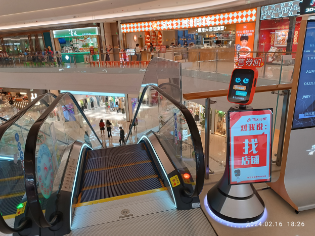 Eine Rolltreppe in einem Einkaufszentrum mit Menschen, die hinuntergehen, ein Bildschirm mit Text auf der rechten Seite und verschiedene Geschäfte und Gegenstände im Hintergrund.