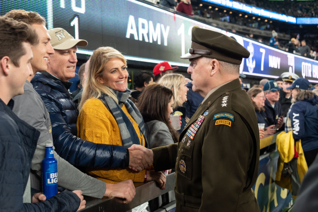 Ein Mann in Milit"aruniform gibt einer Frau die Hand bei einem Fussballspiel, mit einer Menge Menschen im Stadion und beleuchteten Tafeln mit Text und Zahlen im Hintergrund.