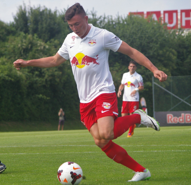 Ein professioneller Fußballspieler schießt einen Ball auf einem Rasenfeld mit Bäumen und einem klaren blauen Himmel im Hintergrund, wobei ein Schild mit der Aufschrift "RB Leipzig" zu sehen ist.