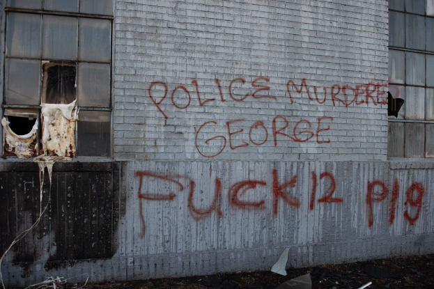 Ein Gebäude mit Graffiti, das "Polizei Mord George Fuck 12 P19" auf der Seite trägt, mit Fenstern und diversen Gegenständen vorne.