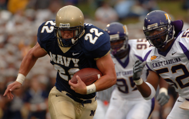 Ein Fußballspieler in einem marineblauen Trikot und Helm rennt mit dem Ball während eines Spiels, der Hintergrund durch ihre Geschwindigkeit unscharf.