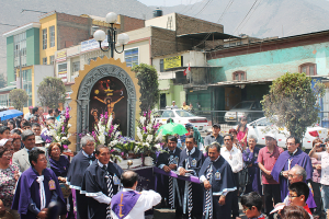 Menschen auf einer Straße vor der Stadt, mit einem Kreuz und Blumensträußen im Vordergrund, Autos im Hintergrund und Gebäuden, Bäumen und Straßenlaternen weiter hinten.