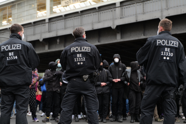 Polizeibeamte in schwarzen Uniformen und Masken stehen vor einer Menge während einer Demonstration, mit einer Brücke und einem Gebäude im Hintergrund.