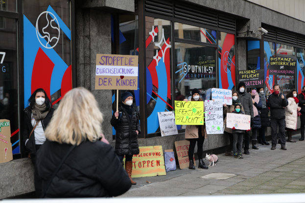 Eine Gruppe maskierter Personen mit Schildern und Protestplakaten vor einem Glasgebäude, mit Kameras, Texttafeln und einem Hund.