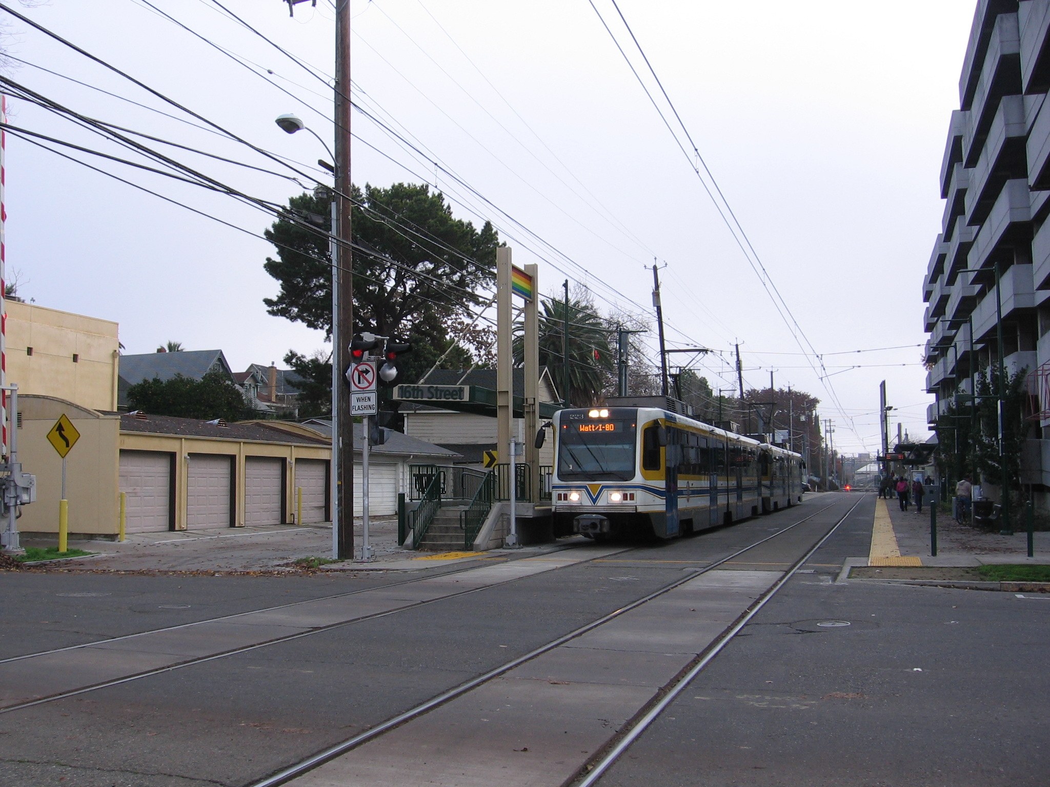 Eine Stadtbahn fährt auf einer Straße mit hohen Gebäuden auf beiden Seiten, Strommasten mit Oberleitungen, Fußgängern auf dem Gehweg, Bäumen im Hintergrund und einem klaren blauen Himmel.