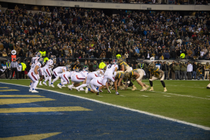 Ein Fußballspiel im Gange mit Spielern in Sportkleidung und Helmen auf dem Feld, Zuschauern, Geländern und Texttafeln im Hintergrund.