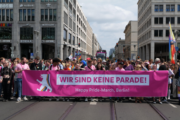 Eine Gruppe von Menschen geht auf einer Straße in Berlin, Deutschland, mit einem pinken Banner, auf dem "Happy Pride March" steht, vorbei an Gebäuden, Laternenmasten und Verkehrszeichen mit Masten und einem bewölkten Himmel im Hintergrund.