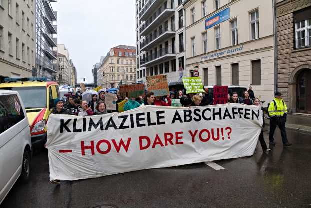 Gruppe von Menschen mit einem Banner "Klimaziele Abschaffen - Wie Wagst Du Es?" auf der Straße unterwegs mit Fahrzeugen, Gebäuden, Schildern und einem klaren blauen Himmel im Hintergrund.