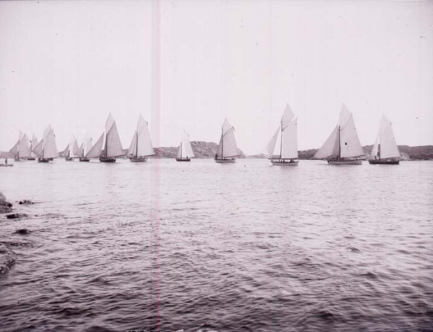 Ein Schwarz-Weiß-Foto mehrerer Segelboote auf dem Wasser mit Bergen im Hintergrund und einem klaren Himmel darüber, mit Text unten, der angibt, dass es ein Foto des New York Yacht Club ist.