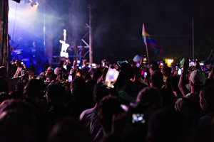 Menge bei einem Konzert mit hochgehaltenen Handys, beleuchtet von Lichtern, umgeben von Pfählen und einer Fahne, während Musiker auf der Bühne spielen.