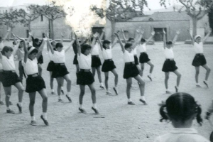 Schwarzes und weißes Bild von jungen Mädchen in Schuluniformen, die auf einer Straße mit Gebäuden und Bäumen tanzen, mit einer Person in Schuhen unten sichtbar.