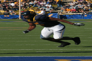 Ein Fußballspieler in voller Montur rennt mit dem Ball auf einem Feld, während Zuschauer auf Stühlen, Geländern, Stufen und einem Pfahl sitzen, mit Bannern im Hintergrund.