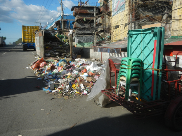 Ein Lastwagen neben einem Müllhaufen auf einer Straße, mit einem Wagen mit Plastikstühlen rechts und Gebäuden, Strommasten, Bäumen und einem bewölkten Himmel im Hintergrund.