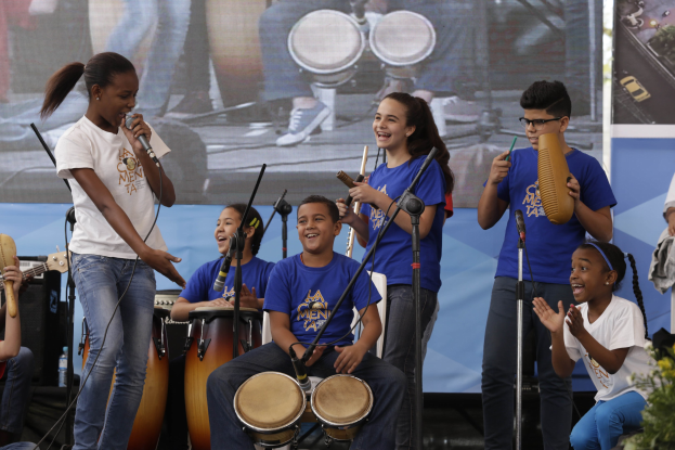 Gruppe von Kindern, die auf einer Bühne Musikinstrumente spielen, mit einer Frau, die ein Mikrofon hält, umgeben von Mikrofonstativen, einem Banner im Hintergrund und Pflanzen auf der rechten Seite.